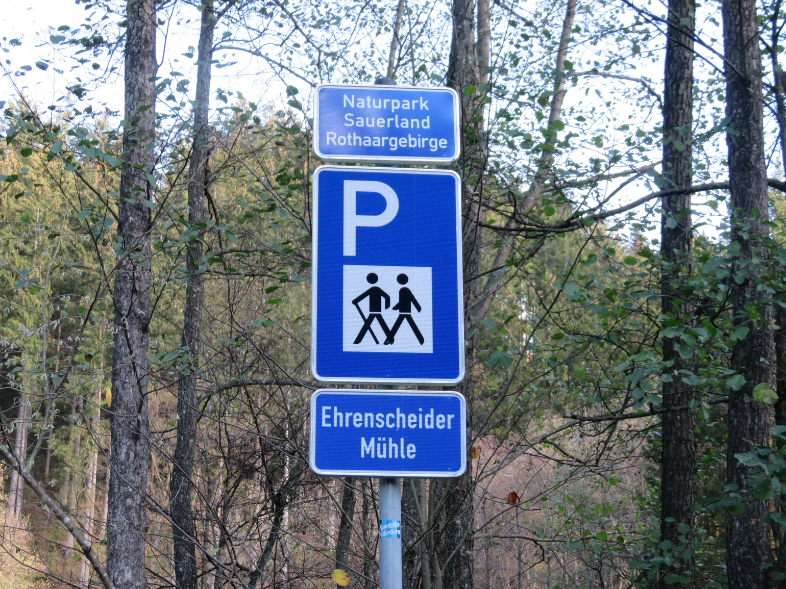 Blaues Schild für einen Wanderparkplatz mit einem weißen P und zwei wandernden Strichmännchen, sowie der Aufschrift Ehrenscheider Mühle. Blaues Schild für einen Wanderparkplatz mit einem weißen P und zwei wandernden Strichmännchen, sowie der Aufschrift Ehrenscheider Mühle.